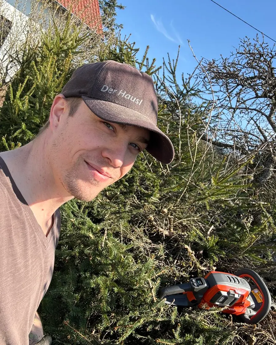 Selfie mit brauner Cap mit Aufschrift Der Hausl vor frisch geschnittenem Fichten-/Heckenschnittgut, orange Akku-Heckenschere daneb
