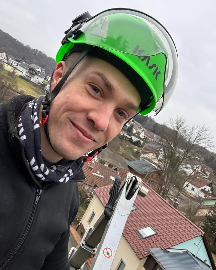 Selfie des Inhabers mit gruenem KASK-Kletterhelm im Hubsteiger-Korb, im Hintergrund Daecher und Haenge des Dorfes.